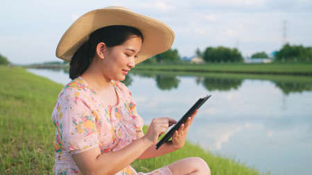 Asian woman relaxing with using portable device sitting on the lawn near the river.