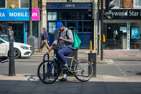 London. UK- 05.18.2021: a young man working as a self employed home delivery rider for the online food ordering company Deliveroo checking on his phone for the next job.のeditorial素材