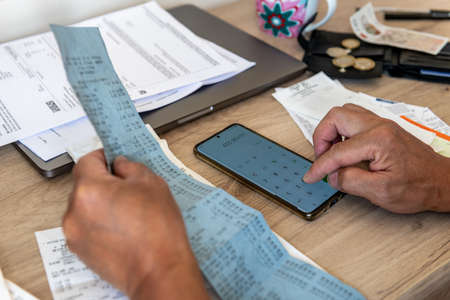 A person calculating the cost of living with a phone calculator and a desk full of bills and till...