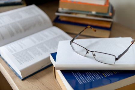 A pair of glasses and on a pile of printed books on a desk.