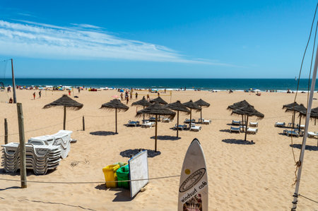 Lisbon. Portugal- 07.08.2023. Surf board, sun beds and umbrellas at Carcavelos Beach with beach sports in the background.のeditorial素材