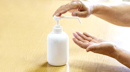 A senior man sprays hands with antiseptic from a plastic spray bottle. Hands with a bottle close-up on a wooden background. Sanitary treatment during an epidemic.の写真素材