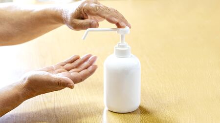 A senior man sprays hands with antiseptic from a plastic spray bottle. Hands with a bottle close-up on a wooden background. Sanitary treatment during an epidemic.の写真素材