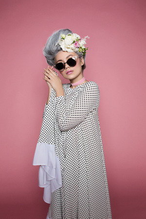 Little beautiful princess in dot dress on soft pink background. Flowers are on head. White-grey hair and sunglasses.の写真素材