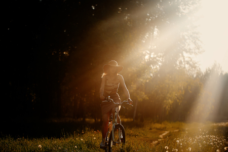 Funny girl driving bicycle outdoor. Sunny summer lifestyle concept. Woman in dress and hat in Field with dandelions. Female ride in park. Light photo effect for text. Copyspace for designの写真素材