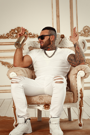 Sexy closeup portrait of Elegant handsome male model with fashion tattoo and a black beard sitting in chair in white casual clothes in hotel interior with gold decor. Professional Studio image.の写真素材