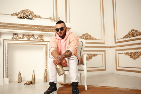 Sexy closeup portrait of Elegant handsome male model with fashion tattoo and a black beard sitting in chair in white casual clothes in hotel interior with gold decor. Professional Studio image.の写真素材