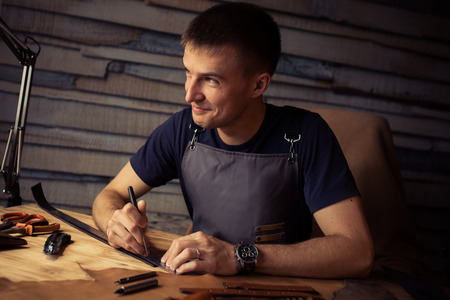 Working process of the leather belt in the leather workshop. Man holding crafting tool and working. Tanner in old tannery. Wooden table background. Close up man arm.の写真素材