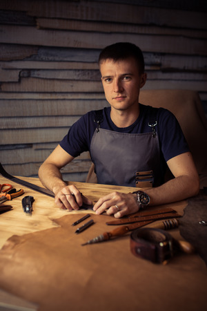 Working process of the leather belt in the leather workshop. Man holding crafting tool and working. Tanner in old tannery. Wooden table background. Close up man arm.の写真素材