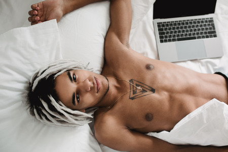 Portrait of handsome african black man with dreadlocks lying with notebook in bed at home. Contrast black and white.の写真素材