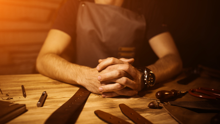 Working process of the leather belt in the workshop.Man holding hands on wooden table.Crafting tools background. Tanner in old tannery.Interlock fingers.Warm Light for text and design.Web banner size.の写真素材