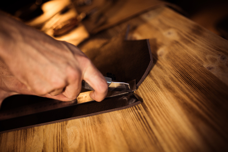 Working process of the leather belt in the leather workshop. Man holding crafting tool and working. Tanner in old tannery. Wooden table background. Close up man arm.の写真素材