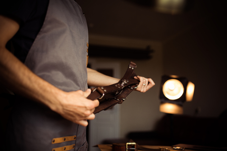 Working process of the leather belt in the leather workshop. Man holding photographer's belt for camera. Tool on wooden background. Tanner in old tannery. Close up master's arm.の写真素材