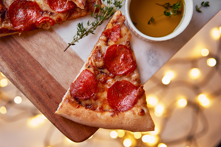 Christmas and New Year atmosphere. Hot Italian pizza with melting tomato, pepperoni and cheese on a white marbel cutting board. Background with lights in bokeh and selective focus.の写真素材