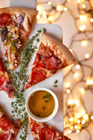 Christmas and New Year atmosphere. Hot Italian pizza with melting tomato, pepperoni and cheese on a white marbel cutting board. Background with lights in bokeh and selective focus.の写真素材