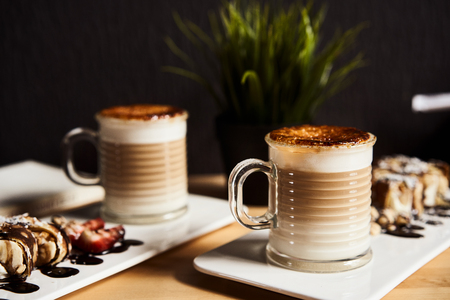 Two cups of Hot latte with baked caramel crust and sweet roll with banana and strawberry on the wooden table in coffe-shop. Coffee concept.の写真素材