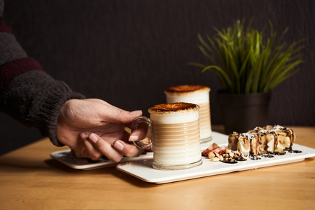 Mans hand hold cup of Hot latte with baked caramel crust and sweet roll with banana and strawberry on the wooden table in coffe-shop. Coffee concept.の写真素材