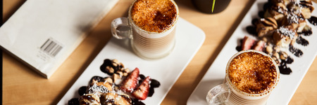 Banner size image of two cups of Hot latte with baked caramel crust and sweet roll with banana and strawberry on the wooden table in coffe-shop. Coffee concept.の写真素材