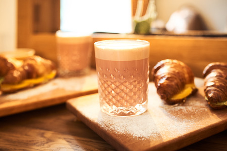 Original authors coffee on beet milk and fresh croissant with fruits on wooden plate on table. Healthy breakfast food concept. Traditional french lunch in morning cafe with gold sunlight flare.の写真素材