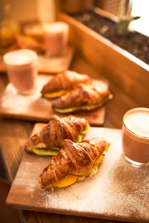 Fresh croissant and original authors coffee on beet milk with fruits on wooden plate on table. Healthy breakfast food concept. Traditional french lunch in morning cafe with gold sunlight flare.の写真素材