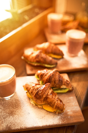 Fresh croissant and original authors coffee on beet milk with fruits on wooden plate on table. Healthy breakfast food concept. Traditional french lunch in morning cafe with gold sunlight flare.の写真素材