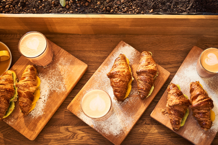 Fresh croissant and original authors coffee on beet milk with fruits on wooden plate on table. Healthy breakfast food concept. Traditional french lunch in morning cafe with gold sunlight flare.の写真素材