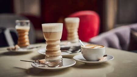 Latte glass with layered latte, cappuccino or mocha with foam on table in cafe with milk saucer and spoon. Three cups of coffee. Food and drink toning image. Web banner size 16 in 9 crop.の写真素材