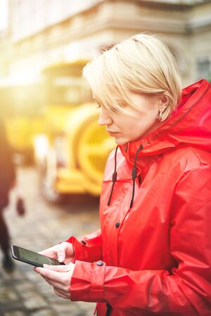 Girl use map on the phone on the street. Portrait of pretty smiling young woman in red bright raincoat searching the place. Sunny day in city. Image with flare.の写真素材