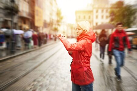 Time concept. Girl looking on watch on the street. Portrait of pretty smiling young woman in red bright raincoat. Sunny raining day in city. Image with yellow flare.の写真素材