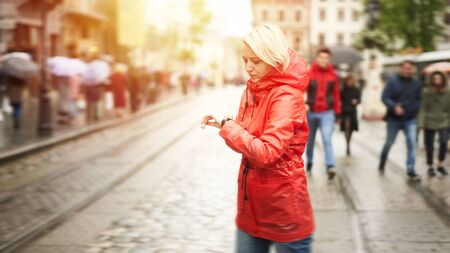 Time concept. Girl looking on watch and waiting friend on the street. Portrait of pretty smiling young woman in red bright raincoat. Web 16 in 9 crop banner size image with flare.の写真素材