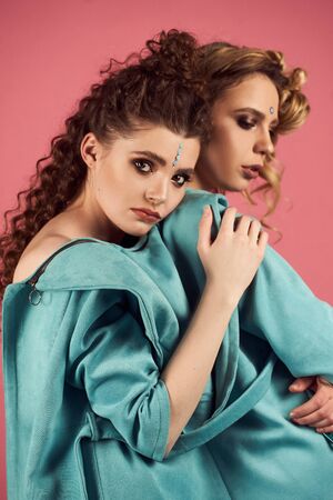 Close up indoor studio fashion portrait of gorgeous women in stylish blue clothes. Blonde and brunette girls on mint-pink background. Head shot of glad curly models.の写真素材