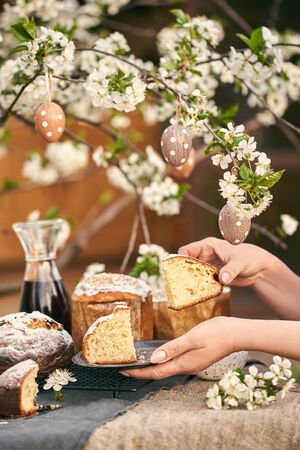 Traditional russian Easter cottage cheese dessert. Woman hands take a piece of orthodox paskha. Kulich cakes on table. Flowers bokeh background.の写真素材