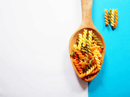 It's time for pasta dish, top view picture of multicolored uncooked fusilli consist of yellow, orange and green on wooden spoon above white and blue  background.の写真素材