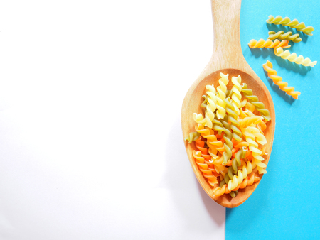 It's time for pasta dish, top view picture of multicolored uncooked fusilli consist of yellow, orange and green on wooden spoon above white and blue  background.の写真素材