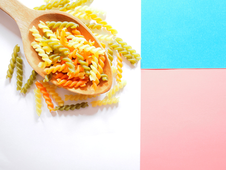 It's time for pasta dish, top view picture of multicolored uncooked fusilli consist of yellow, orange and green on wooden spoon above white, blue and pink  background.の写真素材