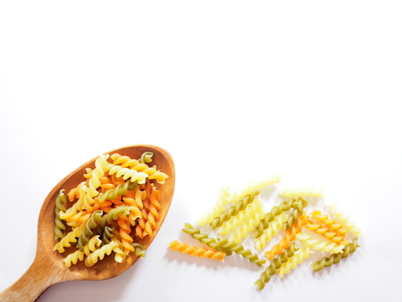 It's time for pasta dish, top view picture of multicolored uncooked fusilli consist of yellow, orange and green on wooden spoon above  white background.の写真素材