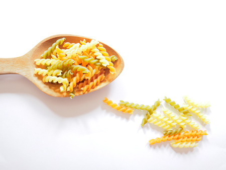 It's time for pasta dish, top view picture of multicolored uncooked fusilli consist of yellow, orange and green on wooden spoon above  white background.の写真素材