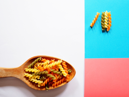 It's time for pasta dish, top view picture of multicolored uncooked fusilli consist of yellow, orange and green on wooden spoon above white, blue and pink  background.の写真素材