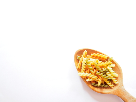 It's time for pasta dish, top view picture of multicolored uncooked fusilli consist of yellow, orange and green on wooden spoon above  white background.の写真素材