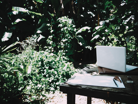 Businesses happen everywhere, just have inspiration. The picture of white laptop, paper, pencil, watch and glasses on green forest background in the afternoon.  selective focusの写真素材