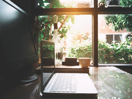 Businesses happen everywhere, just have inspiration. The picture of white laptop and a cup of coffee on wooden table near the window at home office. selective focusの写真素材