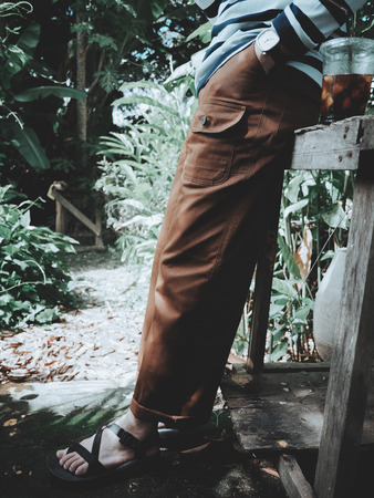 Portrait of a man wearing brown pants with striped shirt and black sandals with iced coffee on green forest background.の写真素材