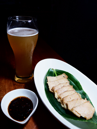 A sliced of grilled pork on white dish with Thai spicy sauce and a glass of beer above wooden table.の写真素材