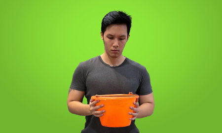 Asian Person looking down into orange bucket, wearing dark gray t-shirt isolated on bright green screen backdropの写真素材