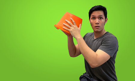 Asian Person holding orange bucket with expressive face, wearing dark gray t-shirt isolated on bright green screen backdropの写真素材