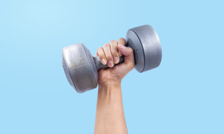 Hand raising dumbbell in the air isolated on blue studio background close up view. Gym exercise, sport activity to build muscleの写真素材