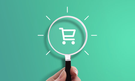 Hand holding magnifying glass with shopping cart icon, symbol of e-commerce, online shopping, and purchaseの写真素材
