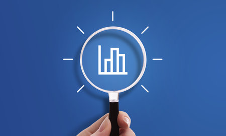 Hand holding magnifying glass with bar graph icon, symbol of data, performance, and analyticsの写真素材