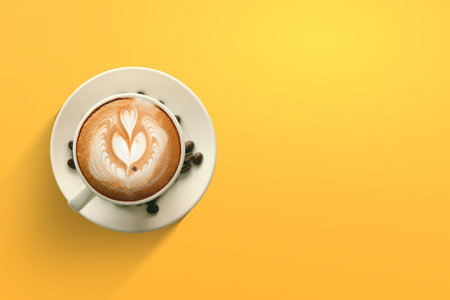 Top view of cup of cappuccino with beautiful latte art on frothy milk, served with coffee beans on a saucer over a bright yellow background.の写真素材