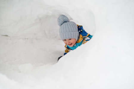 cute young boy child is playing outside in igloo fort tunnel he dug in pile of snow on winter day.の写真素材
